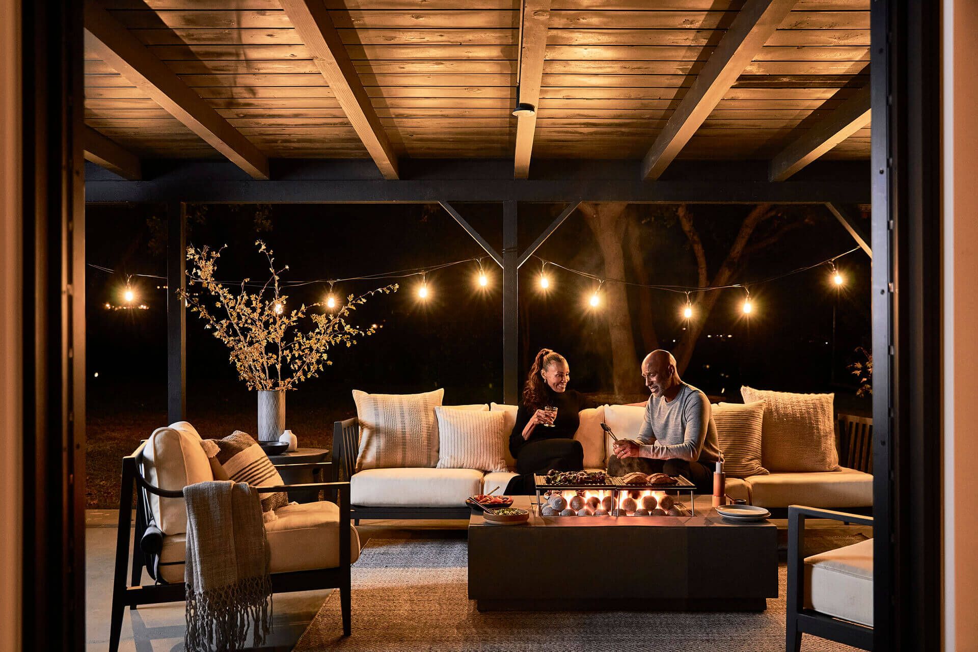 Two people seated beside the lit fire pit table at night, warming themselves by the flame
