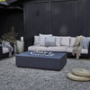Concrete fire pit table lit at dusk on a backyard patio, surrounded by black wicker sofas with sandstone cushions