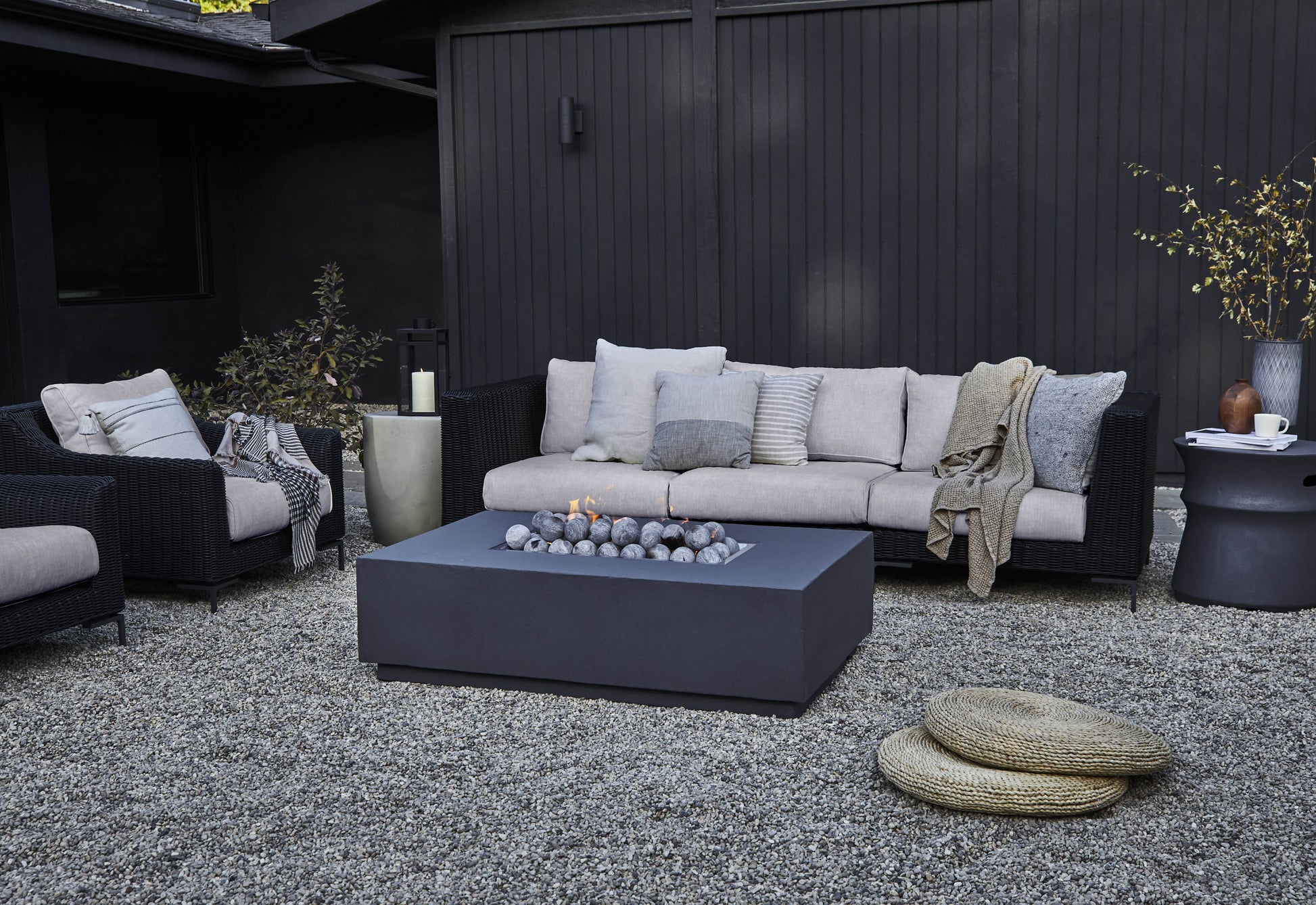 Concrete fire pit table lit at dusk on a backyard patio, surrounded by black wicker sofas with sandstone cushions